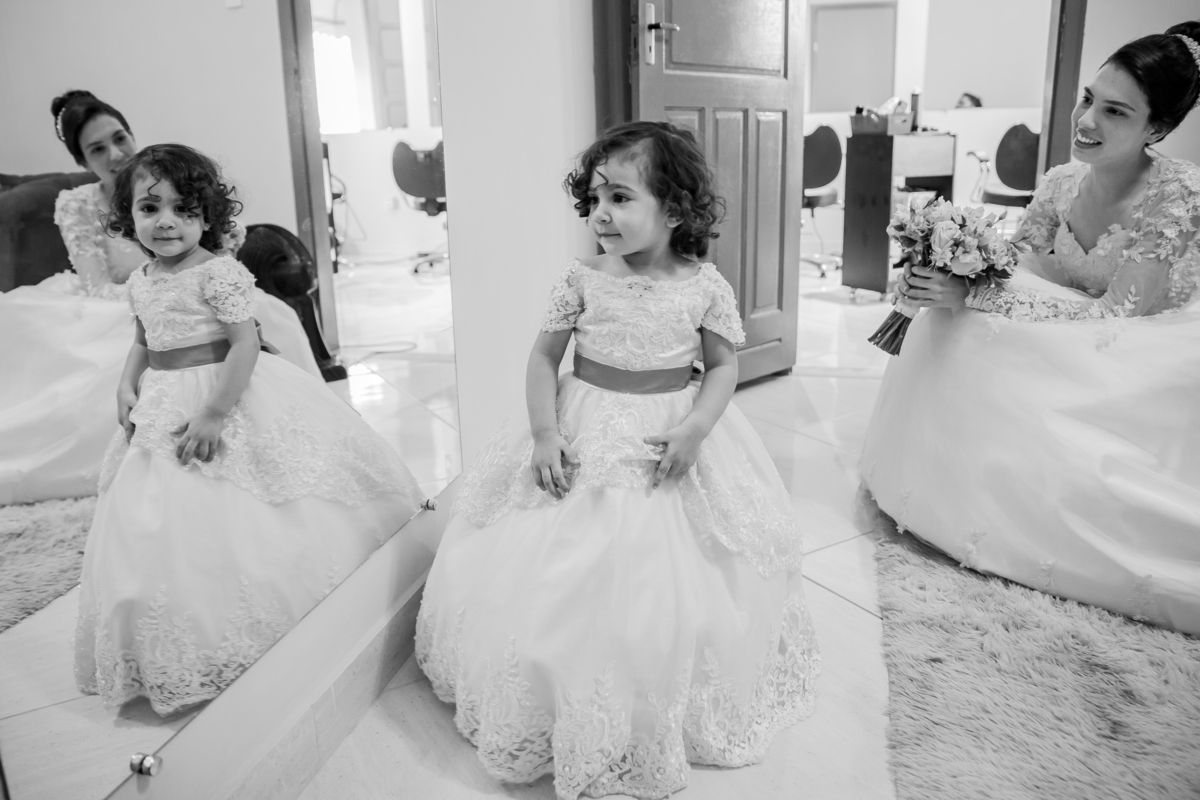 Foto em preta e branco da noiva com seu buquê com flores em tons de rosas com seu vestido lindo com flores em 3D de frente para o espelho olhando a daminha durante making of em salão de beleza em Pirapetinga para casamento em Santo Antônio de Pádua interi