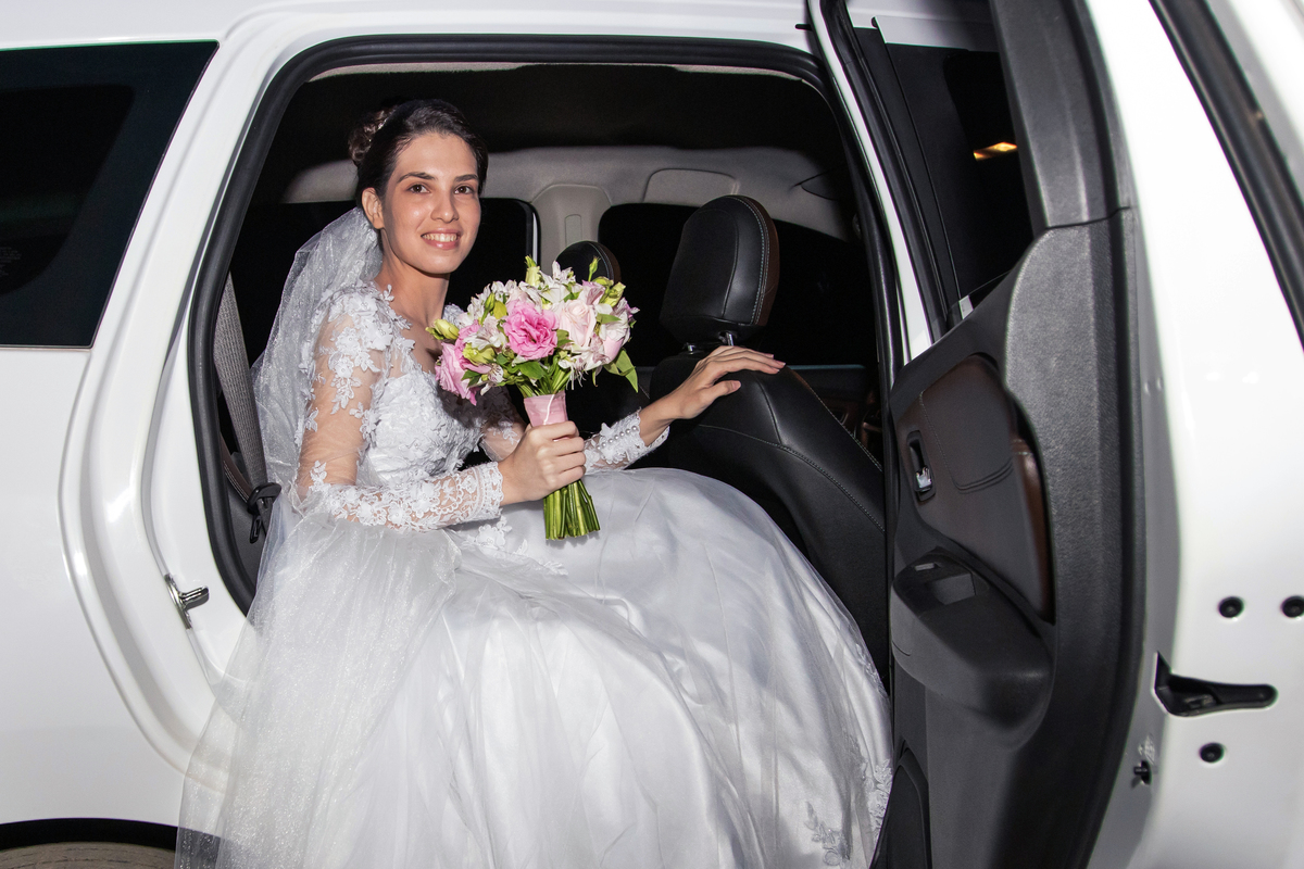 Noiva sentada no carro, segurando seu buquê com flores em tons rosa esperando a hora de entrar durante cerimônia de casamento ao ar livre em Santo Antônio de Pádua interior do Rio de Janeiro.