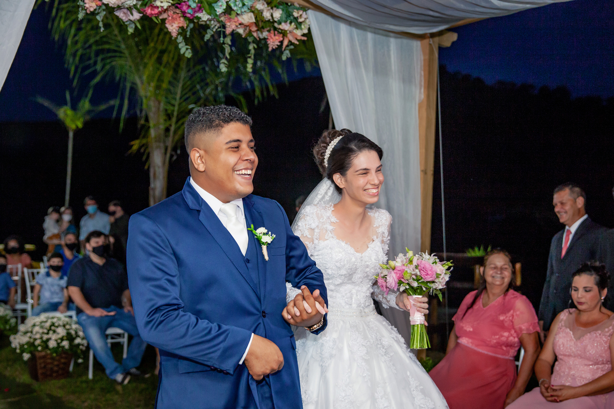 Noivos sorrindo e felizes durante cerimônia de casamento ao ar livre. Noivo com terno azul escuro e noiva com vestido com flores em 3D em Santo Antônio de Pádua interior do Rio de Janeiro.