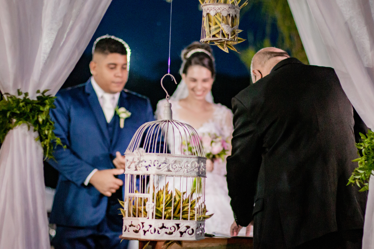 Noivos sorrindo e felizes durante cerimônia de casamento ao ar livre. Noivo com terno azul escuro e noiva com vestido com flores em 3D em Santo Antônio de Pádua interior do Rio de Janeiro.