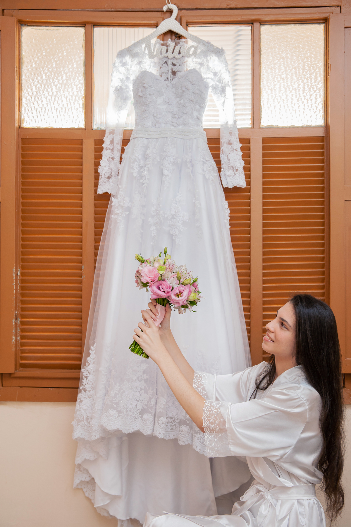 Vestido da noiva pendurado em uma janela de madeira antiga durante Making of noiva em salão de beleza em Pirapetinga, noiva segurando o buquê em tons de rosa para casamento em Santo Antônio de Pádua interior do Rio de Janeiro.