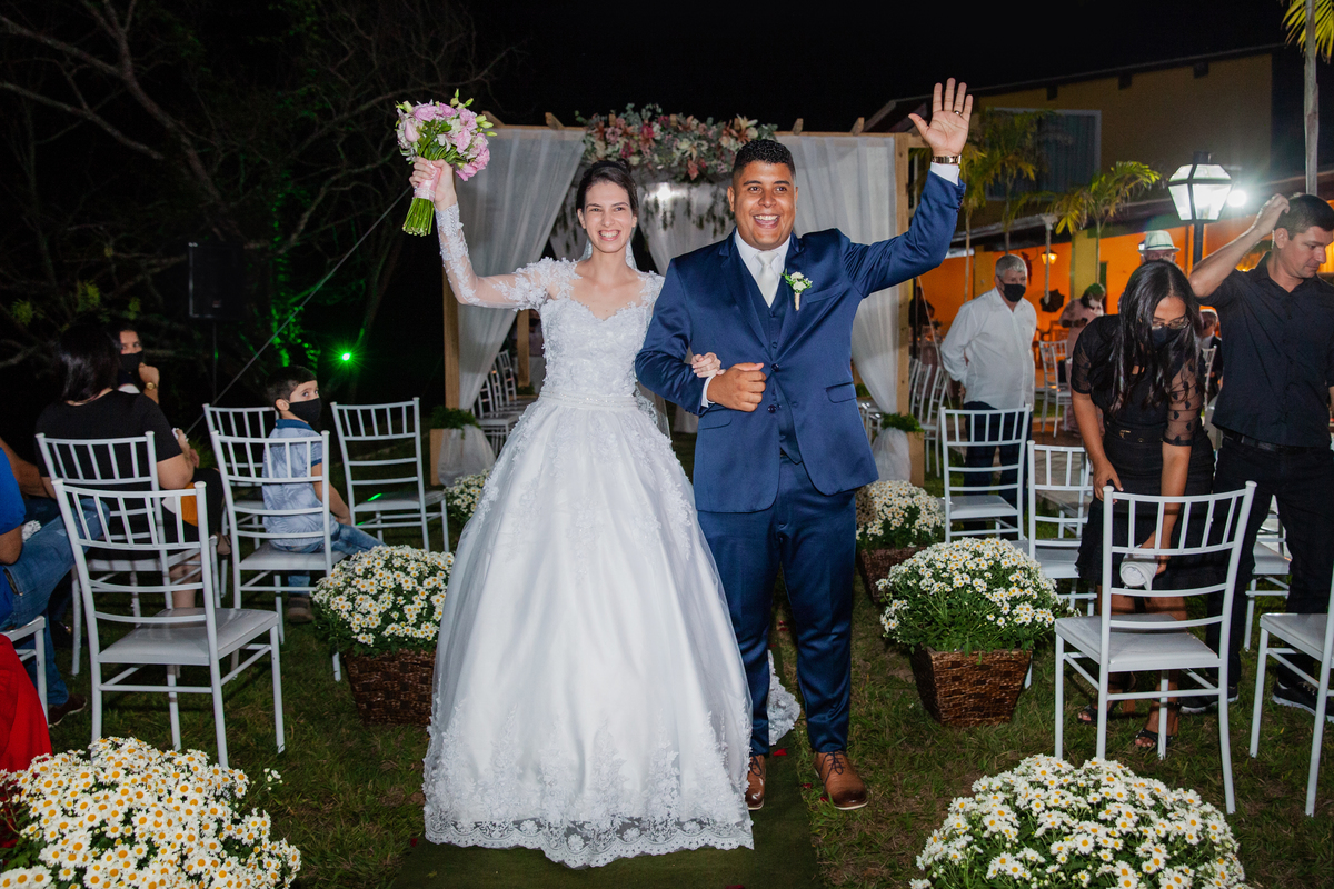 Noivos sorrindo e felizes na saída do casamento ao ar livre. Noivo com terno azul escuro e noiva com vestido com flores em 3D em Santo Antônio de Pádua interior do Rio de Janeiro.
