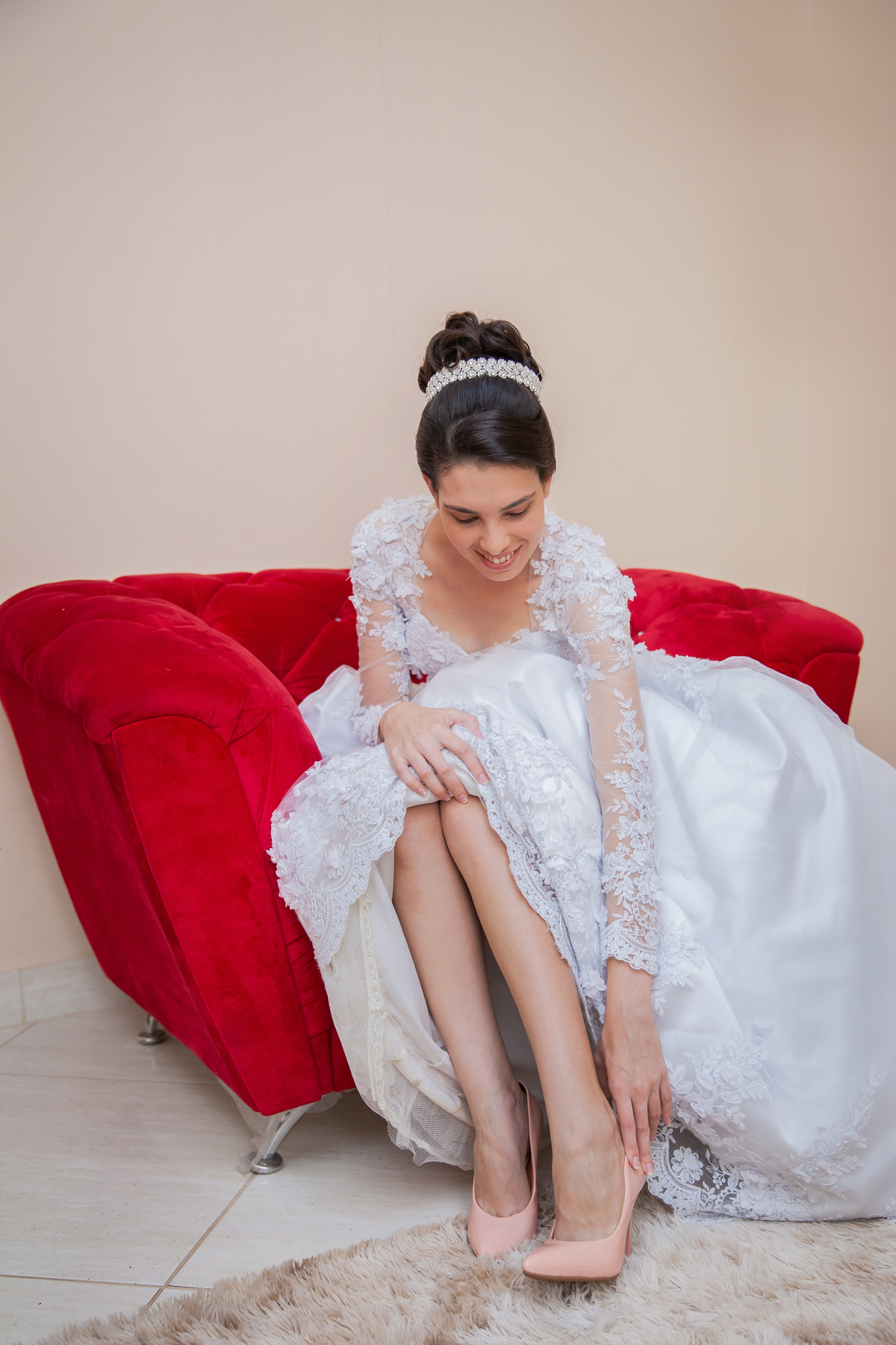 Noiva colocando o sapato com seu vestido lindo com flores em 3D sentada em sofá vermelho vermelho durante making of em salão de beleza em Pirapetinga para casamento em Santo Antônio de Pádua interior do Rio de Janeiro.
