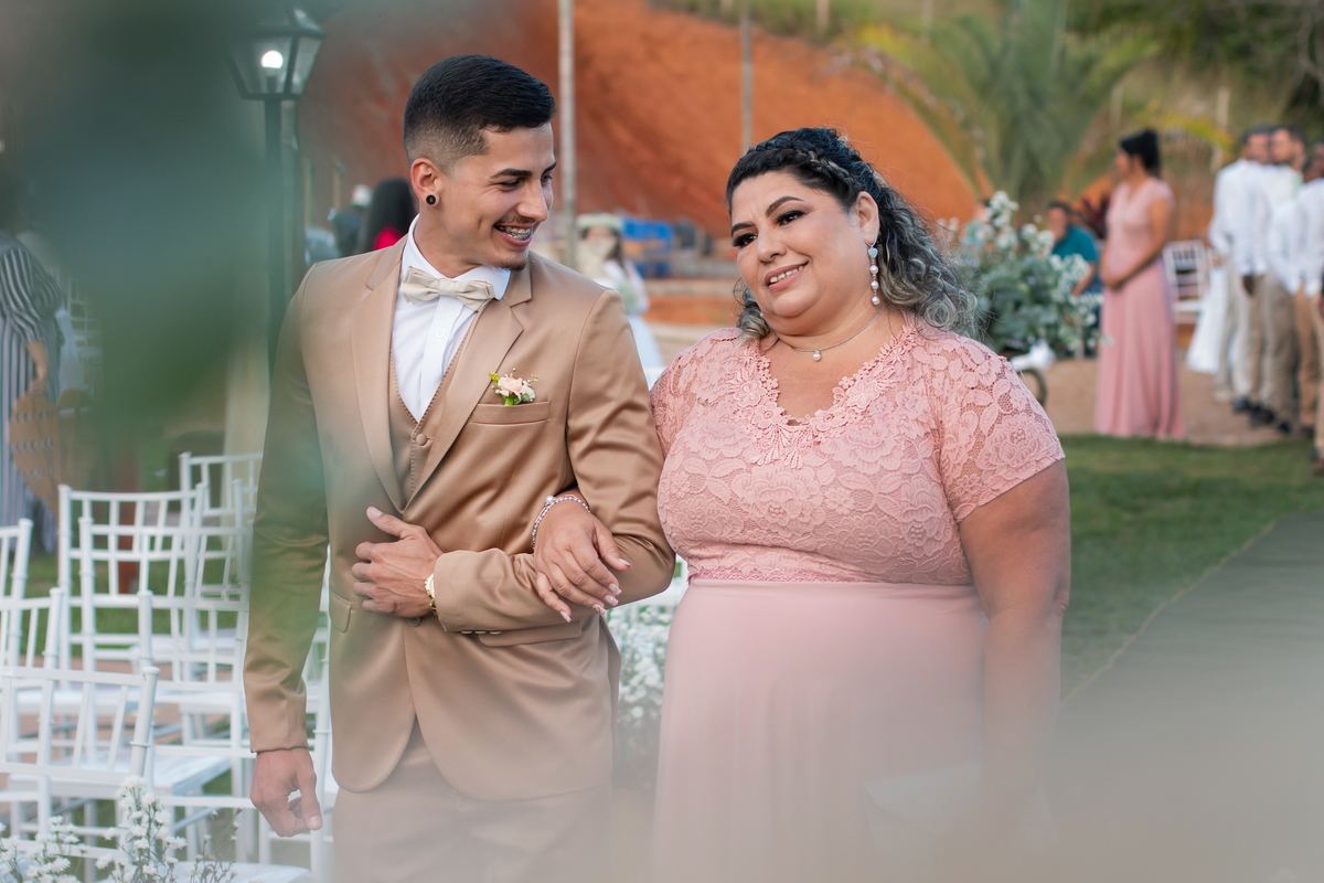 Noivo com terno dourado, feliz, de mãos dadas com sua mãe com vestido rosê na entrada de seu casamento ao ar livre durante o dia com um pôr do sol maravilhoso em Santo Antônio de Pádua no interior do Rio de Janeiro.