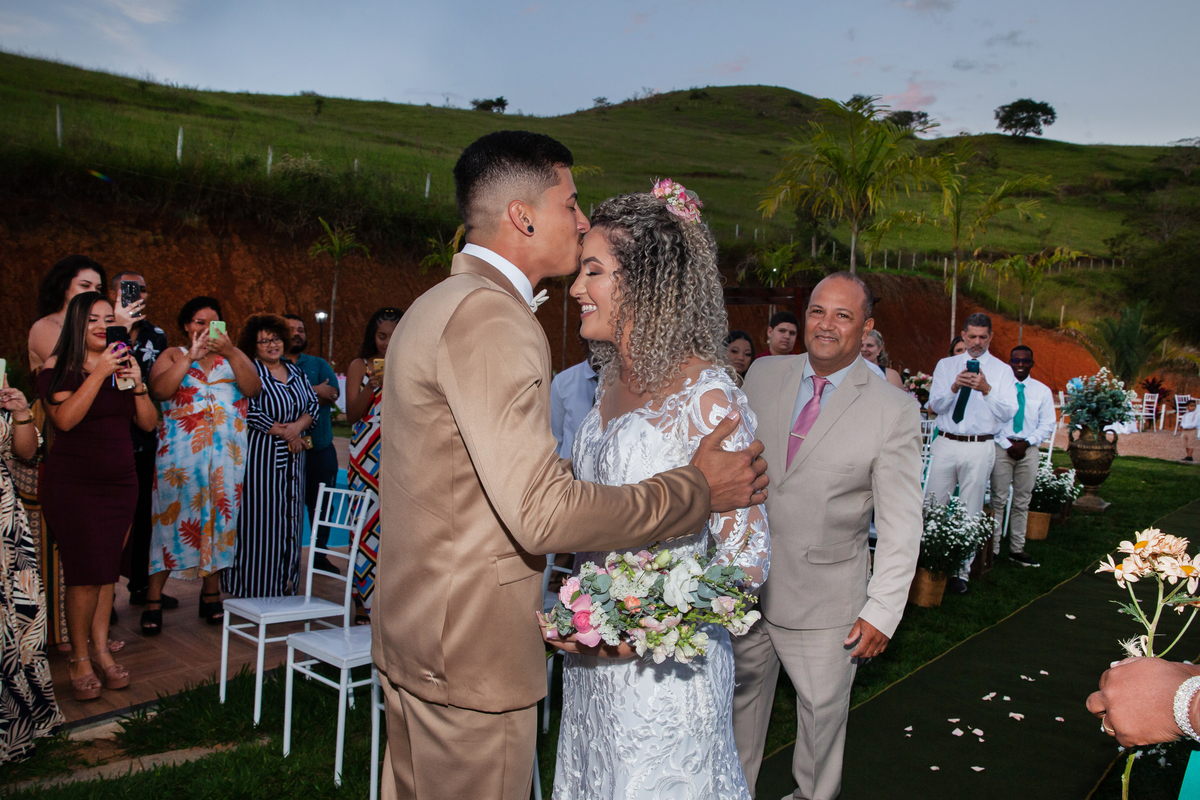 Noivo com terno dourado, beijando a testa da noiva com vestido sereia, na hora da entrada do casamento ao ar livre durante o dia com um pôr do sol maravilhoso em Santo Antônio de Pádua no interior do Rio de Janeiro.