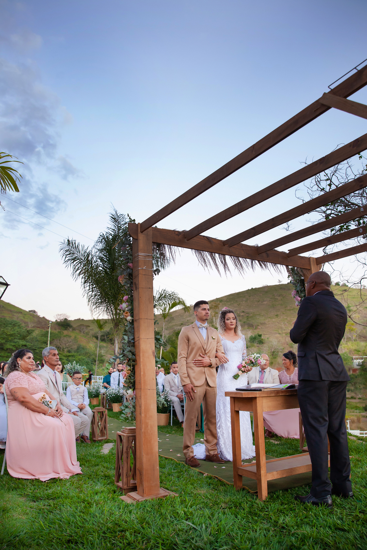 Noivos felizes durante cerimonia de casamento ao ar livre, noiva com vestido sereia e noivo com terno dourado e gravata borboleta. Casamento durante o dia com um pôr do sol maravilhoso em Santo Antônio de Pádua no interior do Rio de Janeiro.