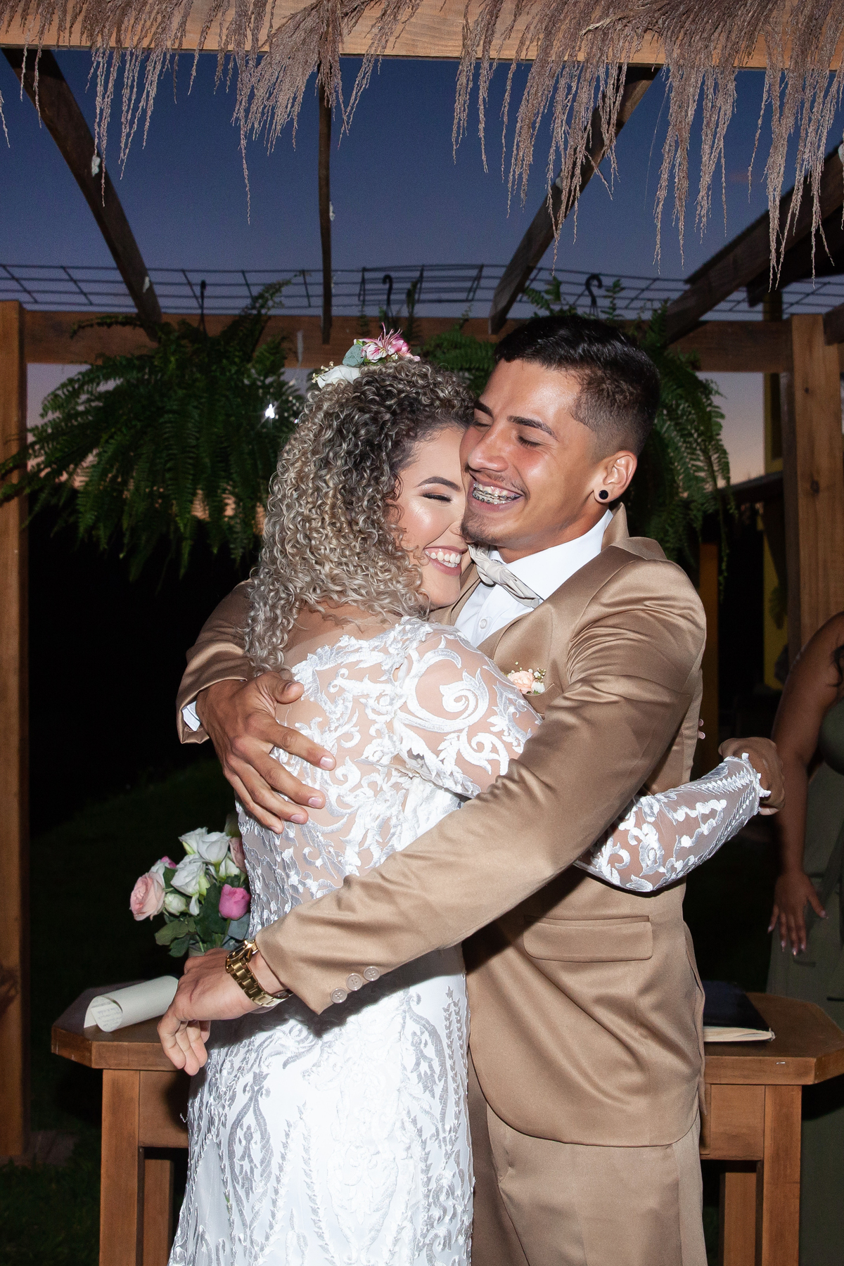 Noivos felizes se abraçando durante cerimonia de casamento ao ar livre, noiva com vestido sereia e noivo com terno dourado e gravata borboleta. Casamento durante o dia com um pôr do sol maravilhoso em Santo Antônio de Pádua no interior do Rio de Janeiro.