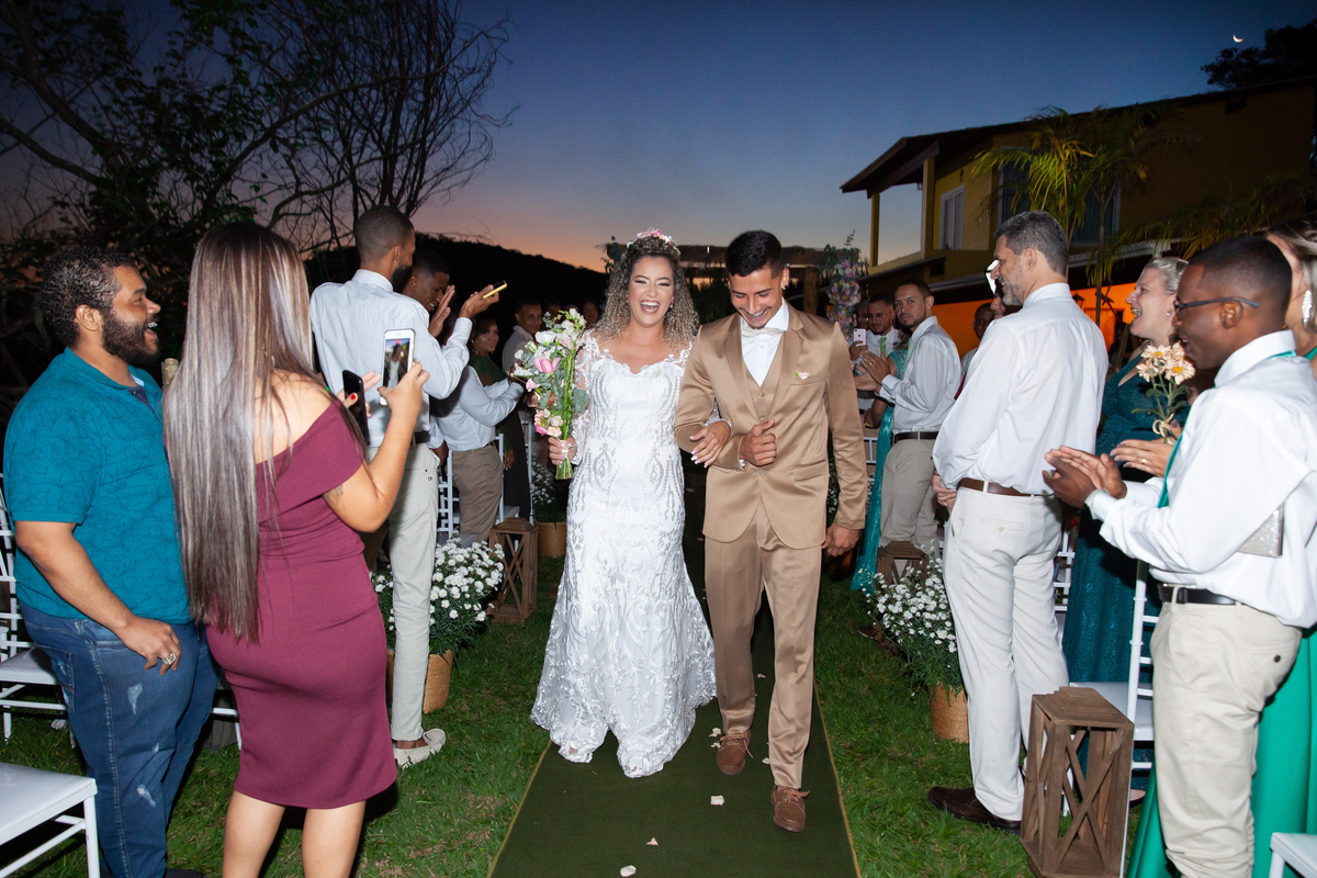 Noivos felizes durante a saída da cerimonia de casamento ao ar livre, noiva com vestido sereia e noivo com terno dourado e gravata borboleta. Casamento durante o dia com um pôr do sol maravilhoso em Santo Antônio de Pádua no interior do Rio de Janeiro.