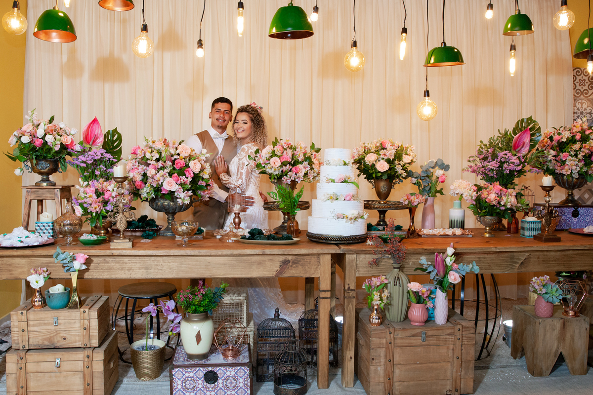 
Noivos se abraçando e felizes no meio da decoração de casamento, com lâmpadas penduradas, moveis rústicos e flores coloridas. Casamento durante o dia ao ar livre  em Santo Antônio de Pádua no interior do Rio de Janeiro.
