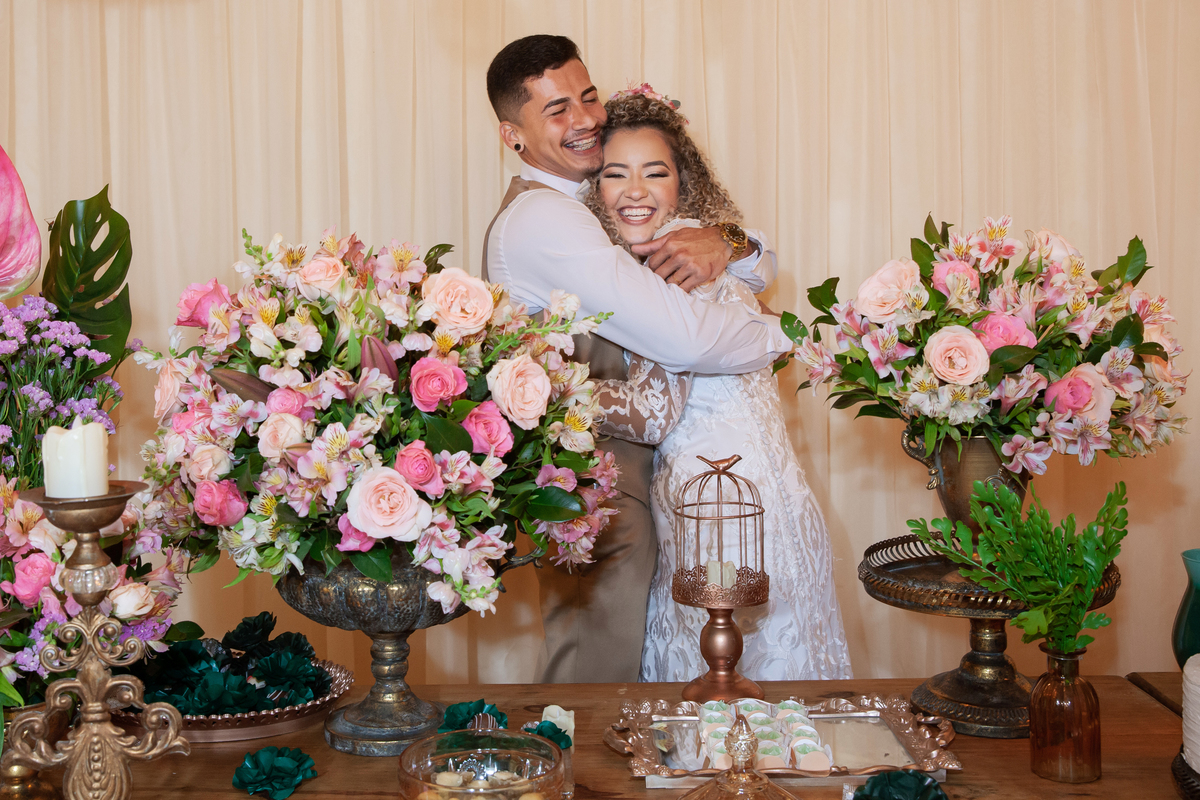 
Noivos se abraçando e felizes no meio da decoração de casamento, com lâmpadas penduradas, moveis rústicos e flores coloridas. Casamento durante o dia ao ar livre  em Santo Antônio de Pádua no interior do Rio de Janeiro.
