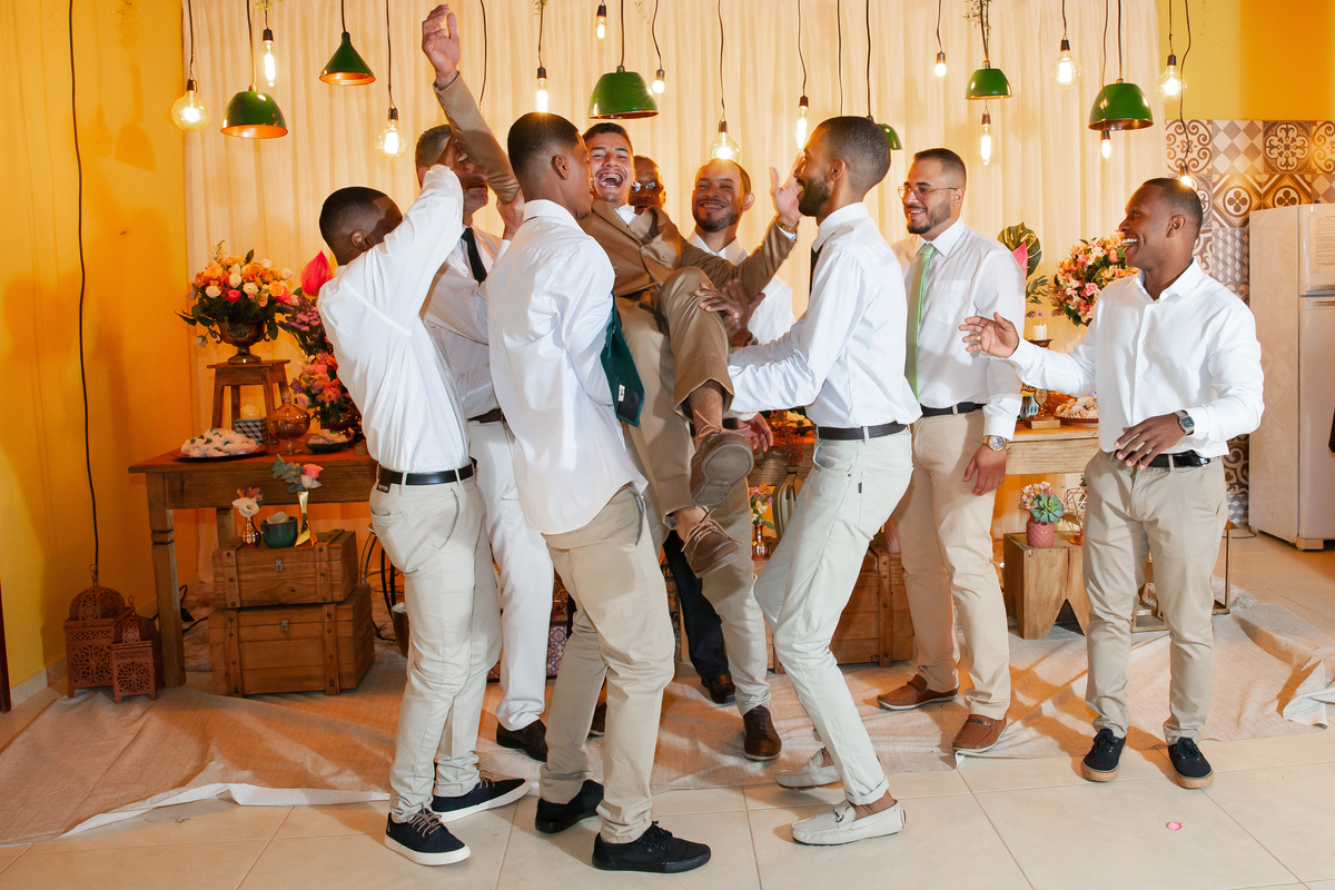 Foto dos Padrinhos de gravata verde pegando o noivo com terno dourado  no colo, todos rindo bastante durante sessão de fotos em Casamento durante o dia ao ar livre  em Santo Antônio de Pádua no interior do Rio de Janeiro.
