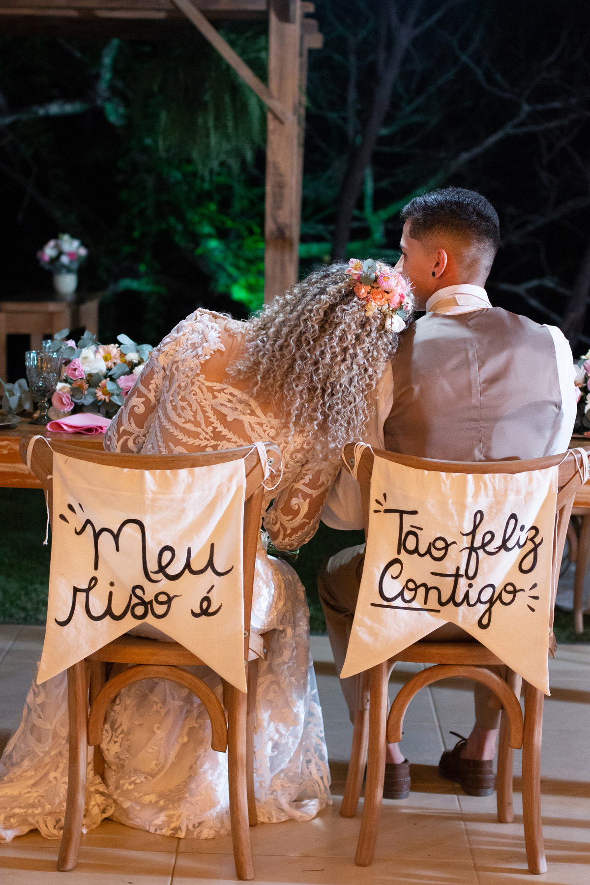 Noiva deitado no ombro do noivo, sentados em cadeiras com plaquinhas escriot: 
