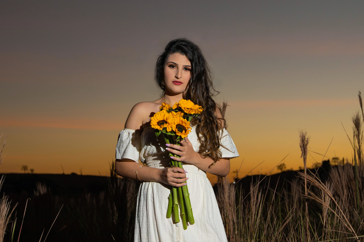 Ensaio de 15 , debutante sorrindo,  com vestido branco em um lindo pôr do sol segurando flores de girassol em Pádua interior do Rio de Janeiro.