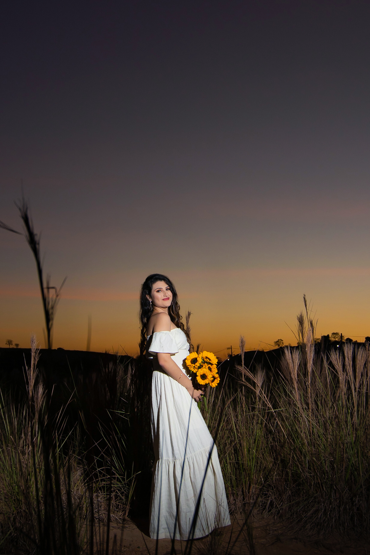 Ensaio de 15 , debutante sorrindo,  com vestido branco em um lindo pôr do sol segurando flores de girassol em Pádua interior do Rio de Janeiro.
