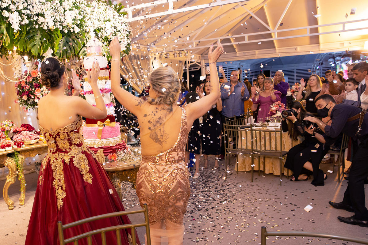 
Debutante linda e feliz com vestido vermelho e dourado cantando parabéns na mesa do bolo de 5 andares em tons de rosa, vermelho, branco e dourado  com chuva de prata em salão de festa maison sainte cecile em Santo Antônio de Pádua.