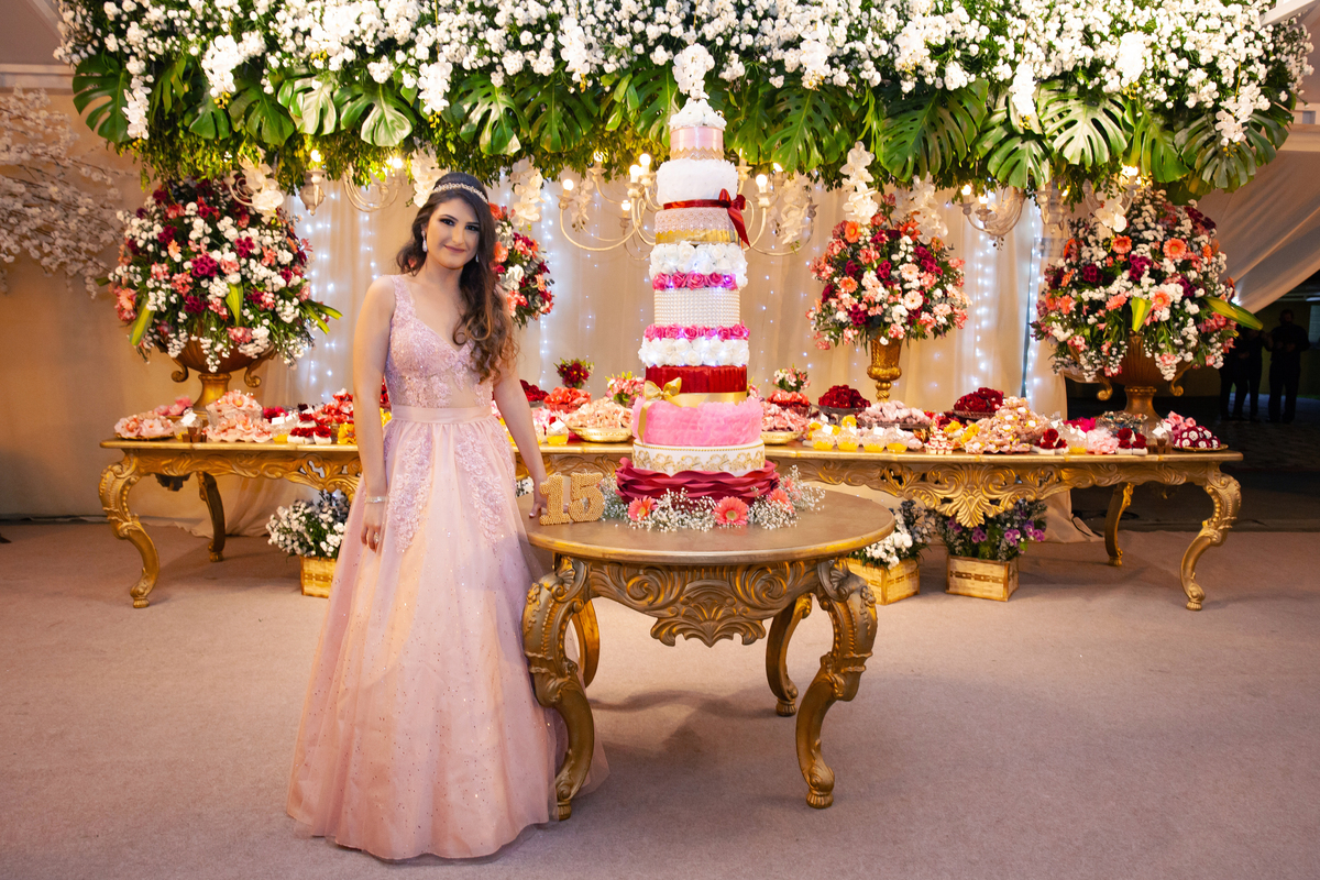 Debutante linda e feliz posando para foto na mesa do bolo de 5 andares em tons de rosa, vermelho, brando e dourado em salão de festa maison saintec ecile em Santo Antônio de Pádua interior do Rio de Janeiro.