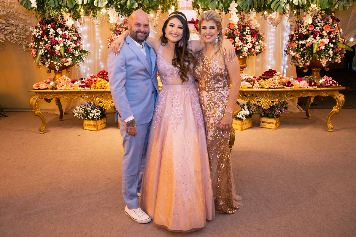 Debutante linda e feliz com vestido rosa e sua família posando para foto na mesa do bolo de 5 andares em tons de rosa, vermelho, brando e dourado em salão de festa maison sainte cecile em Santo Antônio de Pádua interior do Rio de Janeiro.
