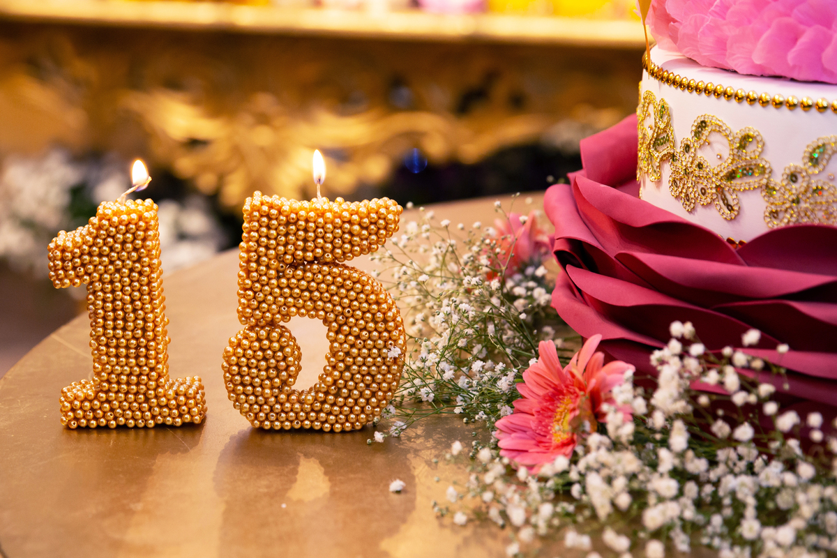 Debutante linda e feliz com vestido vermelho e dourado cantando parabéns na mesa do bolo de 5 andares em tons de rosa, vermelho, branco e dourado  com chuva de prata com vela dourada em salão de festas maison sainte cecile em Pádua.