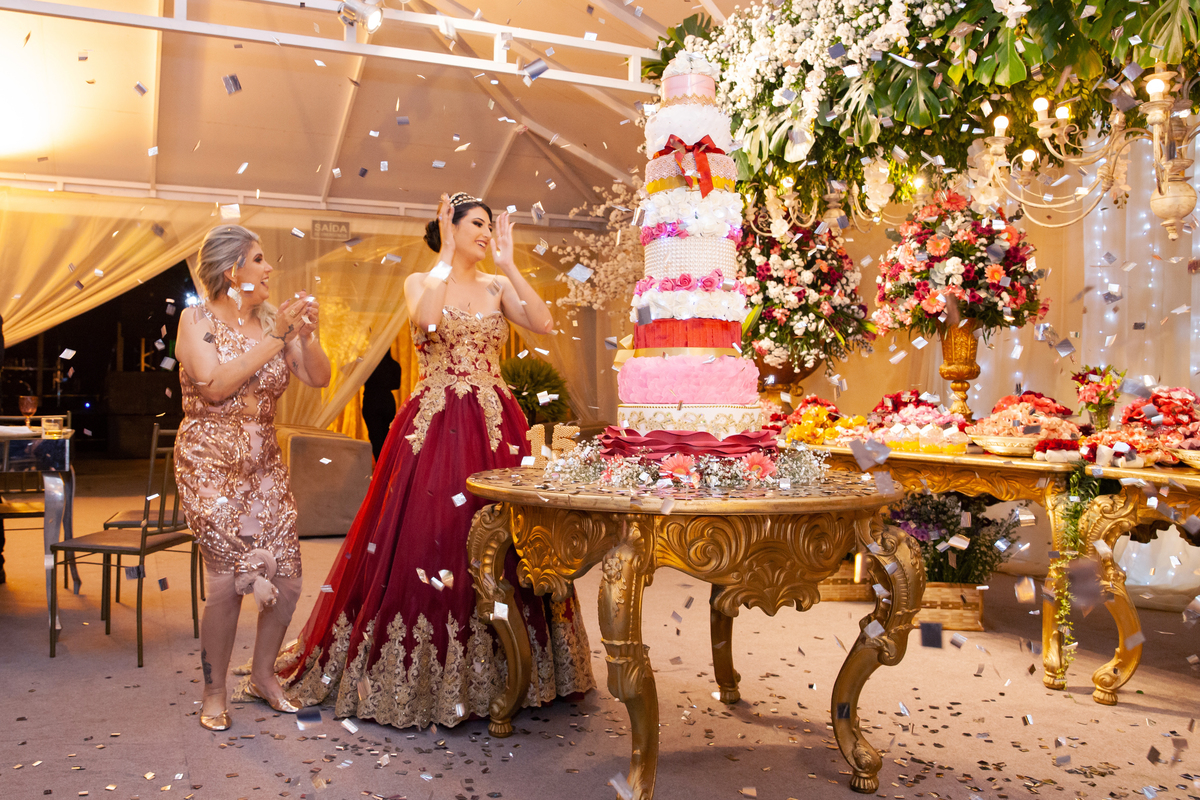 Debutante linda e feliz com vestido vermelho e dourado cantando parabéns na mesa do bolo de 5 andares em tons de rosa, vermelho, branco e dourado  com chuva de prata em salão de festa maison sainte cecile em Santo Antônio de Pádua .