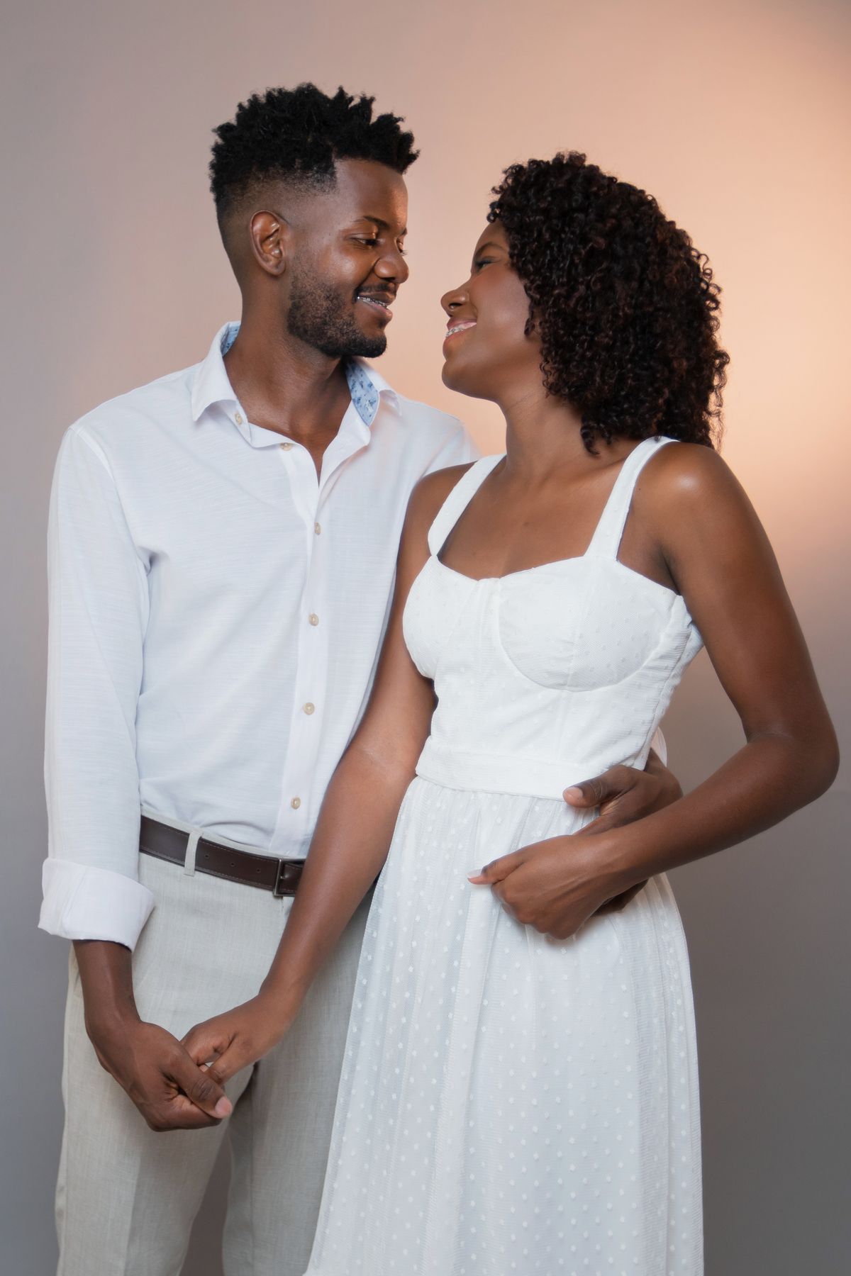 Ensaio pré casamento em estúdio, noiva com vestido branco e noivo com camisa branca  e calça cinza. A noiva está com um buquê de rosas brancas, muito feliz, cabelo trançado e cacheado, em Santo Antônio de Pádua – RJ.