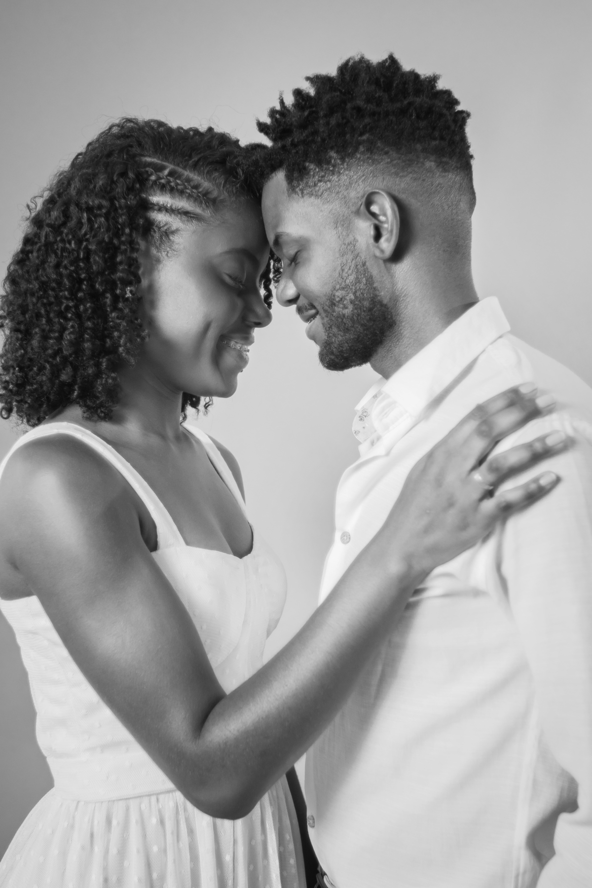 Foto em preto e branco. Ensaio pré casamento em estúdio, noiva com vestido branco e noivo com camisa branca  e calça cinza. A noiva está com um buquê de rosas brancas, muito feliz, cabelo trançado e cacheado, em Santo Antônio de Pádua – RJ.