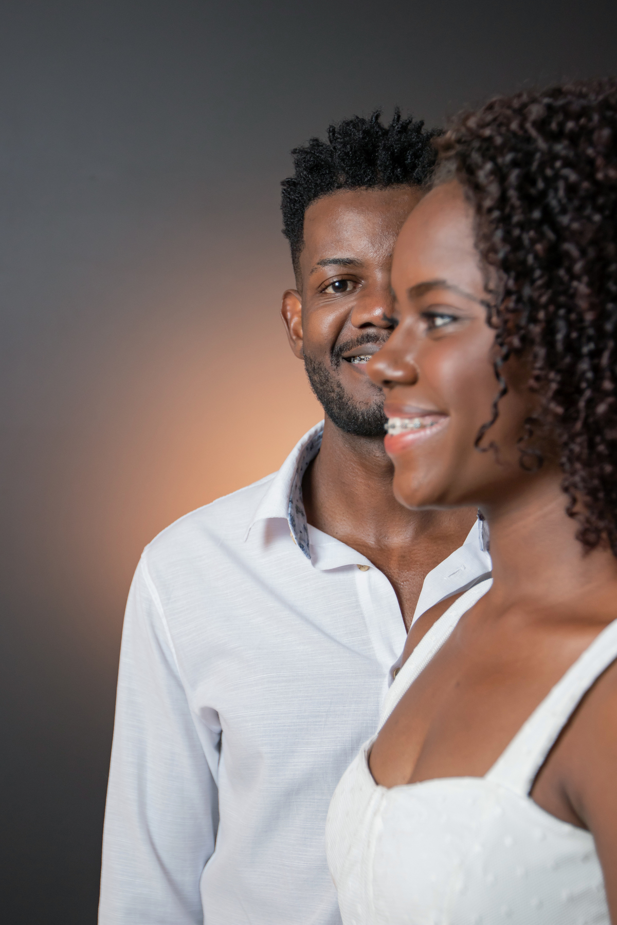 Ensaio pré casamento em estúdio, noiva com vestido branco e noivo com camisa branca  e calça cinza. A noiva está com um buquê de rosas brancas, muito feliz, cabelo trançado e cacheado, em Santo Antônio de Pádua – RJ.