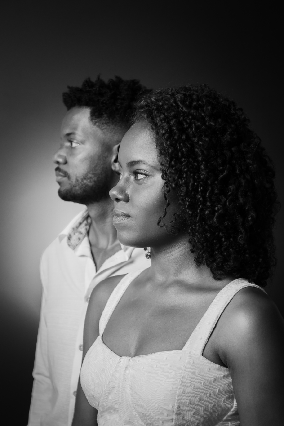 Foto em preto e branco. Ensaio pré casamento em estúdio, noiva com vestido branco e noivo com camisa branca  e calça cinza. A noiva está com um buquê de rosas brancas, muito feliz, cabelo trançado e cacheado, em Santo Antônio de Pádua – RJ.