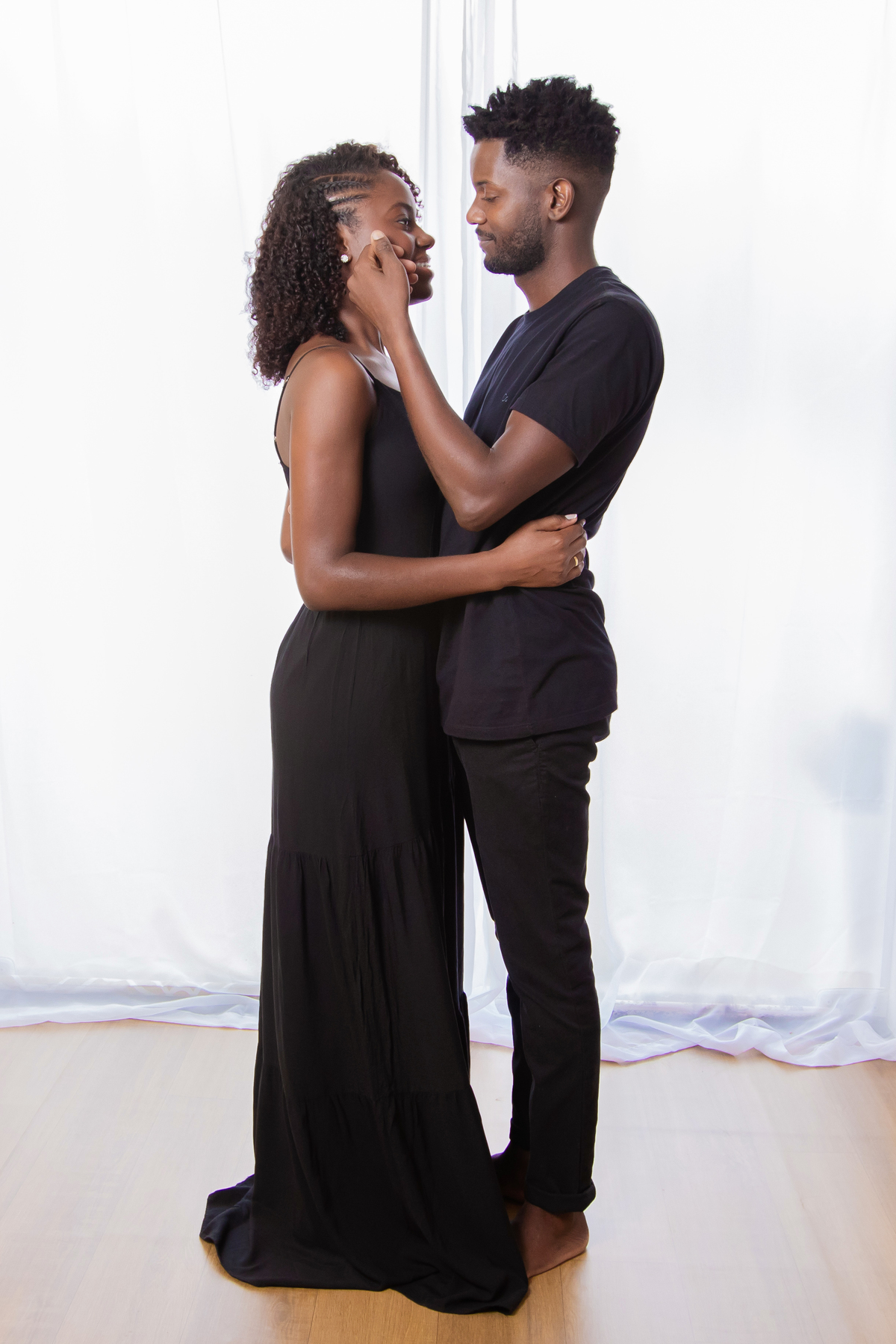 Noiva e noivo com roupas pretas em ensaio pré casamento em estúdio em Santo Antônio de Pádua – RJ. Noiva com buquê de rosas brancas e muito feliz em cenário de flores secas e desidratadas. Noivo fazendo carinho na noiva.