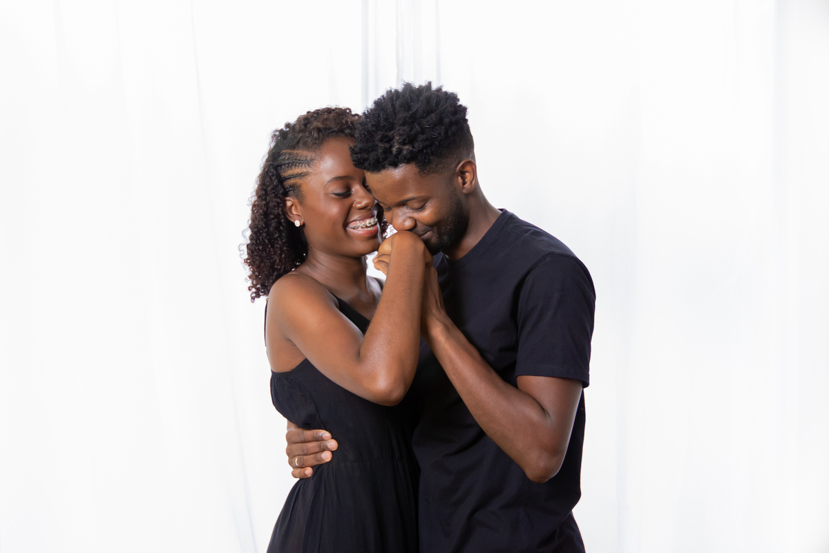 Noiva e noivo com roupas pretas em ensaio pré casamento em estúdio em Santo Antônio de Pádua – RJ. Noiva com buquê de rosas brancas e muito feliz em cenário de flores secas e desidratadas. Noivo beijando a mão da noiva.