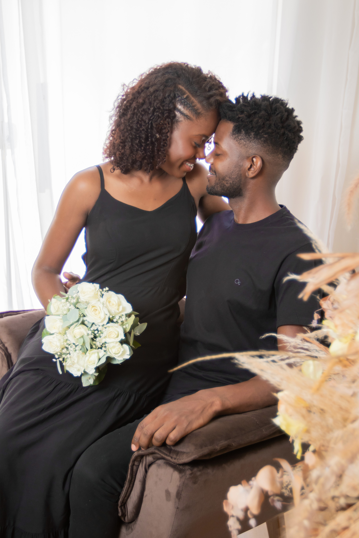 Noiva e noivo com roupas pretas em ensaio pré casamento em estúdio em Santo Antônio de Pádua – RJ. Noiva com buquê de rosas brancas e muito feliz em cenário de flores secas e desidratadas. 