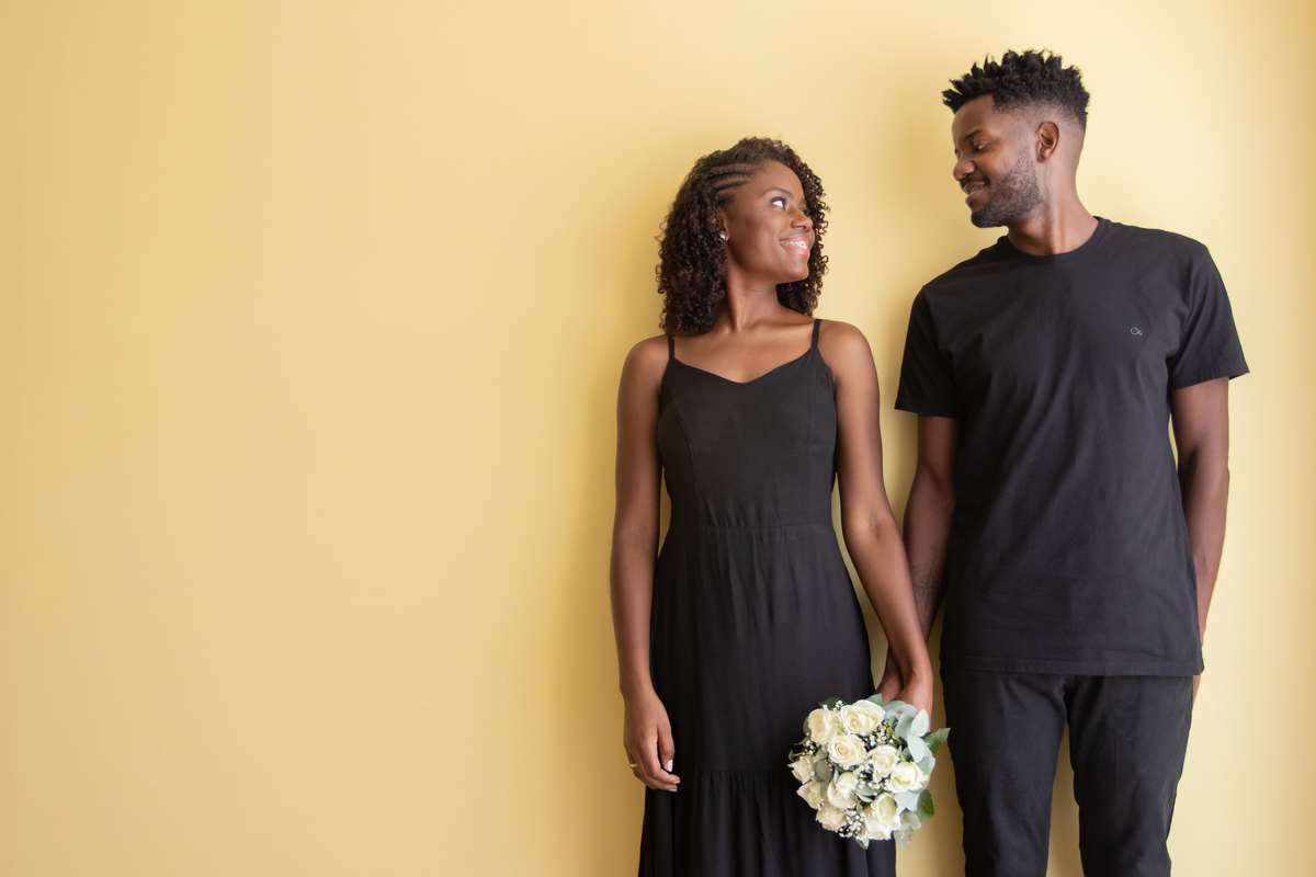 Noiva e noivo com roupas pretas em ensaio pré casamento em estúdio em Santo Antônio de Pádua – RJ. Noiva com buquê de rosas brancas e muito feliz em cenário de flores secas e desidratadas. 