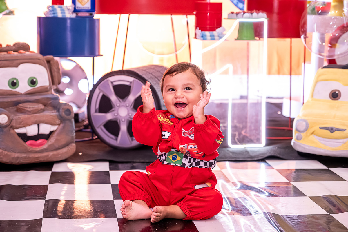 Levi sentado no chão, cercado por elementos da decoração Carros, com um sorriso espontâneo durante a festa.
