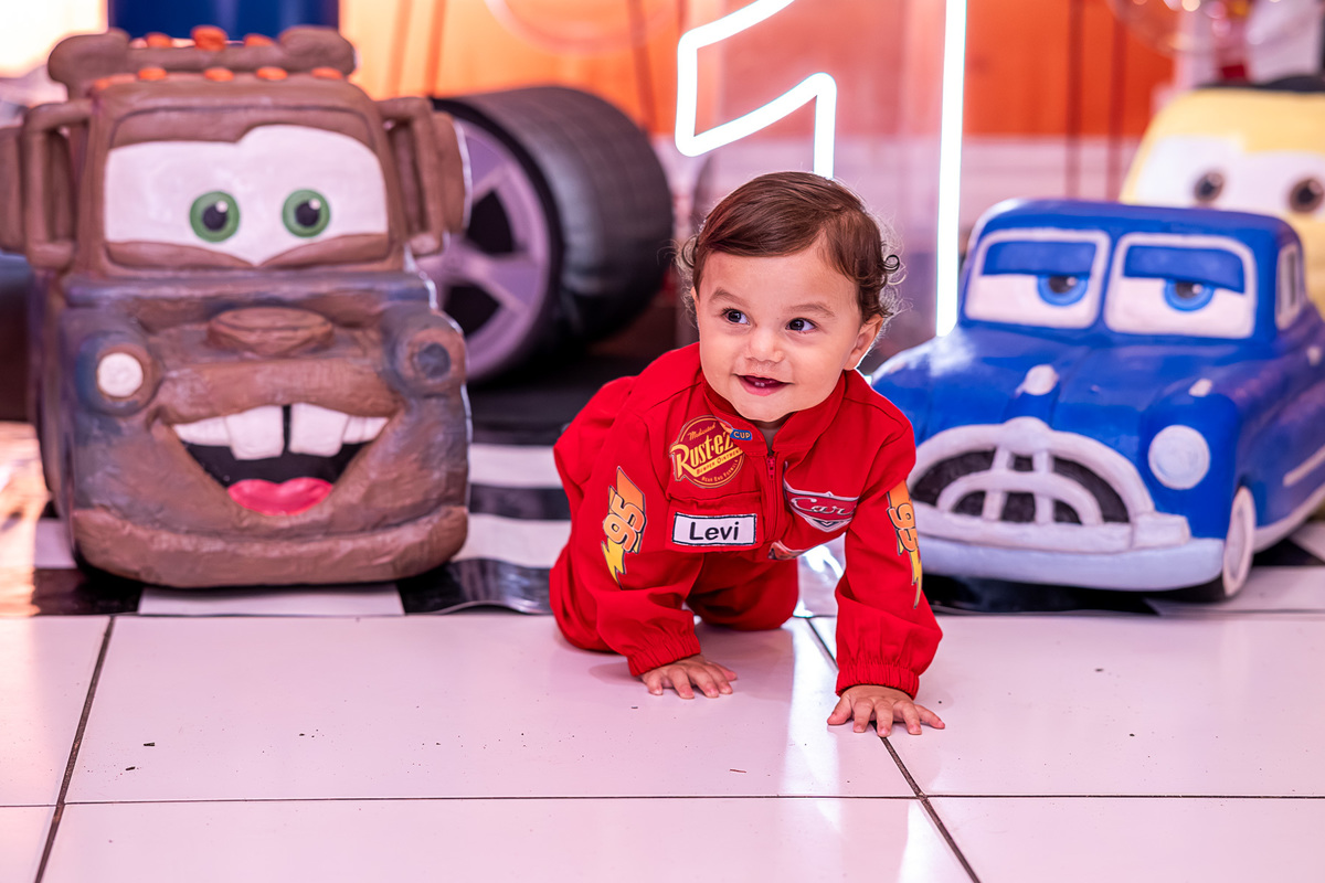 Levi sorrindo e brincando sozinho junto ao painel decorativo do tema Carros na sua festa de 1 ano.