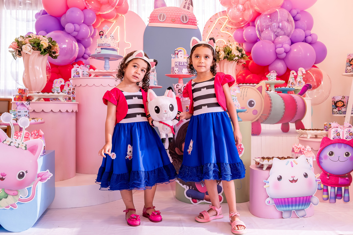 Helena e Heloísa de mãos dadas, caminhando pela festa, com a decoração ao fundo.