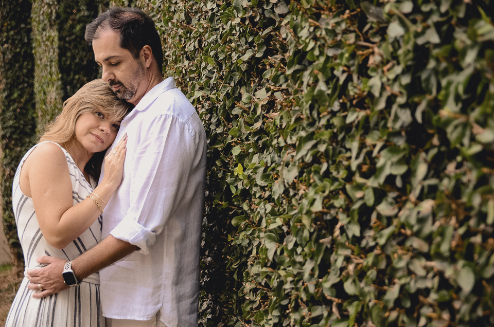 casal está próximo a uma parede forrada de vegetação, ela apoiada com rosto e a mão no peito dele