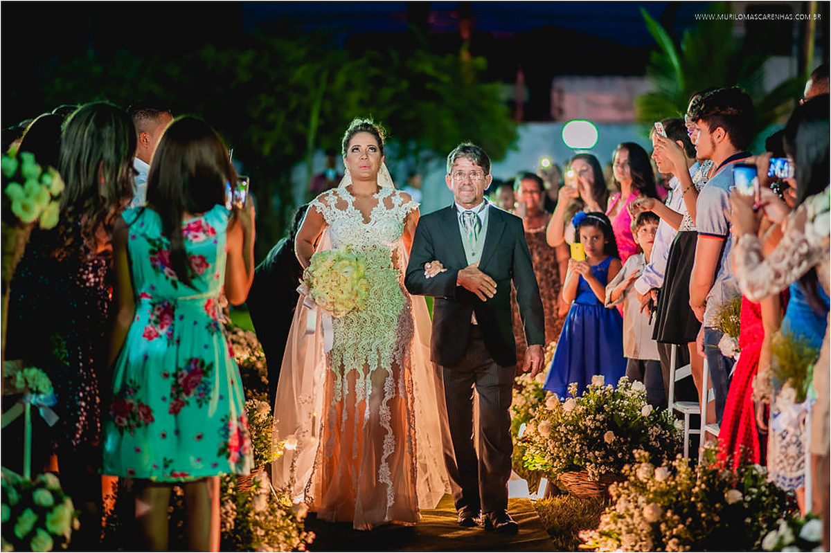 Noiva entra respirando fundo, ao lado do seu pai, enquanto é observada pelos convidados no corredor da igreja