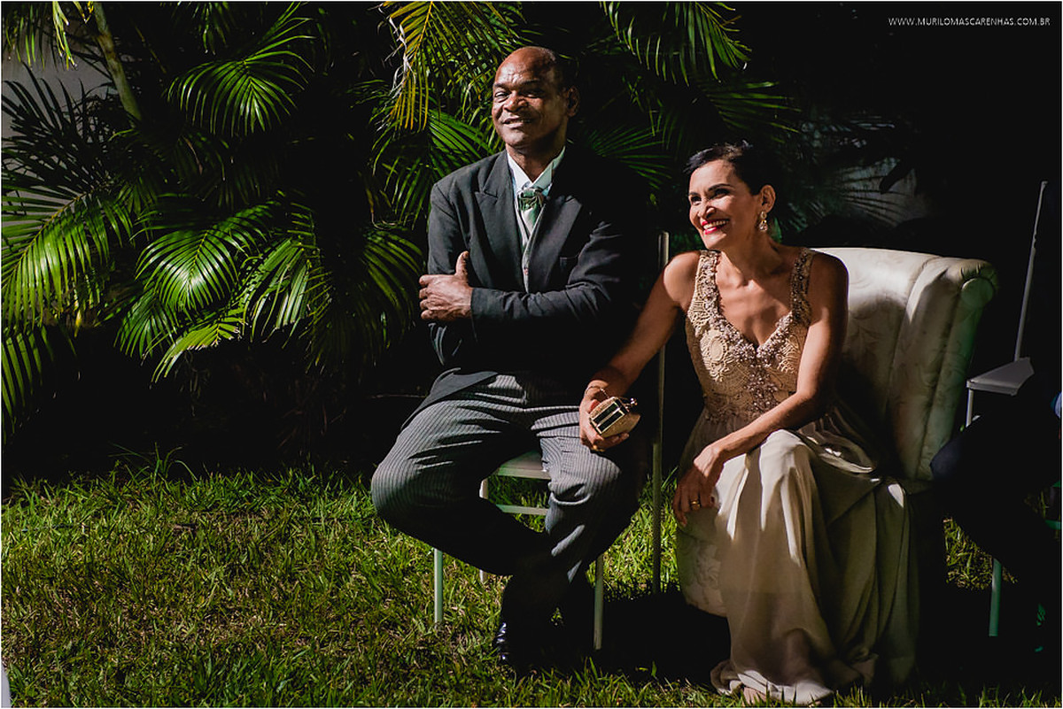 Pai e mãe do noivo, sentados, assistem à cerimônia e sorriem