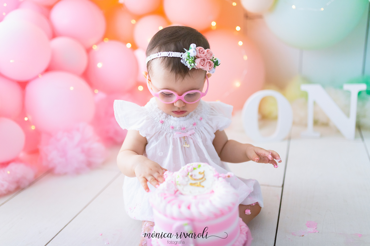 No cenário de Smash the Cake, uma das bebês está sentada olhando para o bolo que está na sua frente e colocando a mão nele. 
Fotografado por Mônicas Fotografia em Maringá-PR