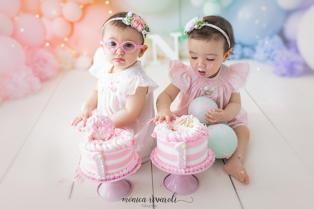 No cenário de Smash The Cake, as bebês estão sentadas com os bolos. As duas estão lambuzadas de chantilly e com a mão no bolo, sentindo a textura.
Fotografado por Mônicas Fotografia em Maringá-PR
