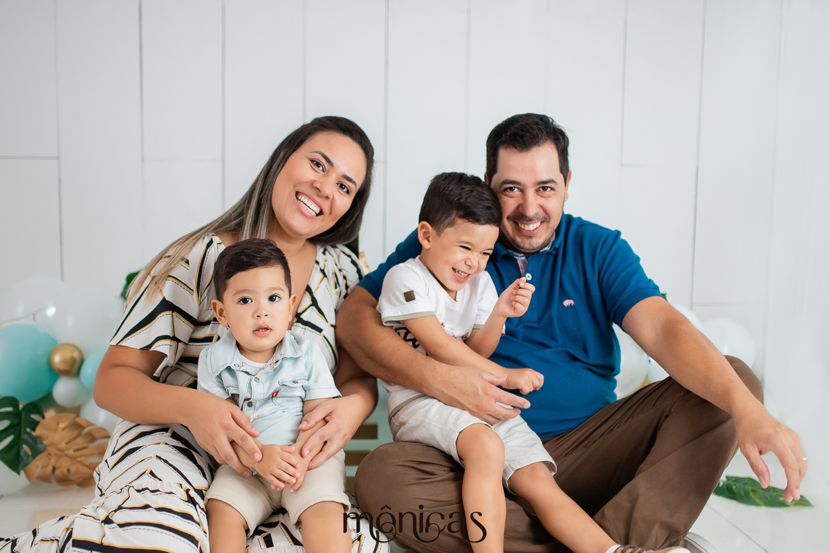 A família está sentada no chão e no fundo está o cenário de Smash. O filho mais novo está no colo da mãe e o filho mais velho no colo do pai. A mãe, o pai e o irmão mais velho estão sorrindo.
Fotografado por Mônicas Fotografia em Maringá-PR