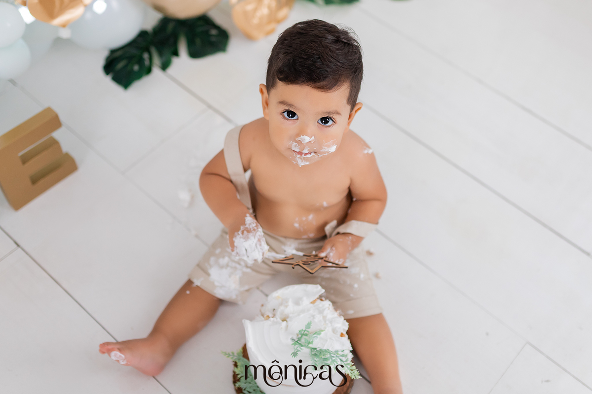 A foto foi tirada de cima, o bebê está olhando para a câmera. O bolo está na frente do bebê e as mãos estão sujas de bolo. Ele está com chantilly no rosto.
Fotografado por Mônicas Fotografia em Maringá-PR