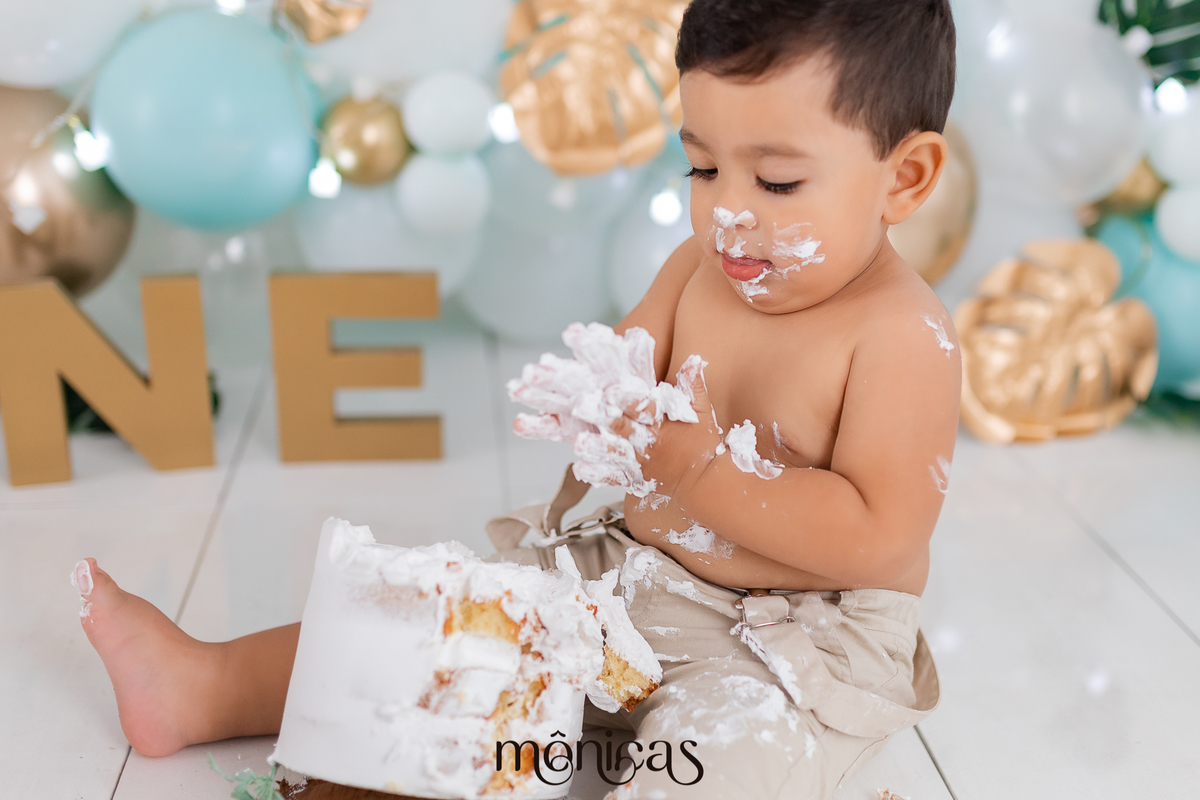 No cenário de Smash, o bebê Caio está olhando para as mãos cheias de chantilly. O bolo está na frente dele.
Fotografado por Mônicas Fotografia em Maringá-PR
