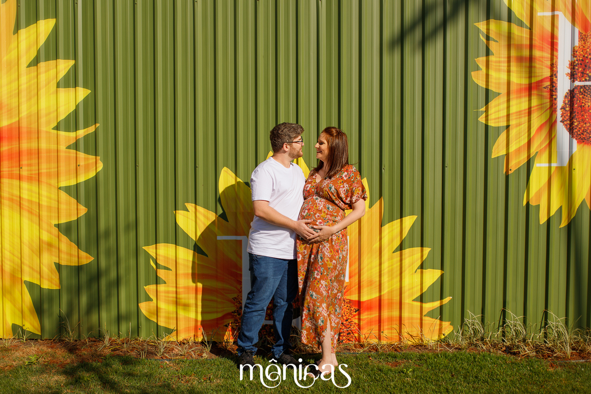 Em frente a um tapume pintados com girassóis, o casal está um de rente para o outro com as mãos na barriga e se olham olho no olho