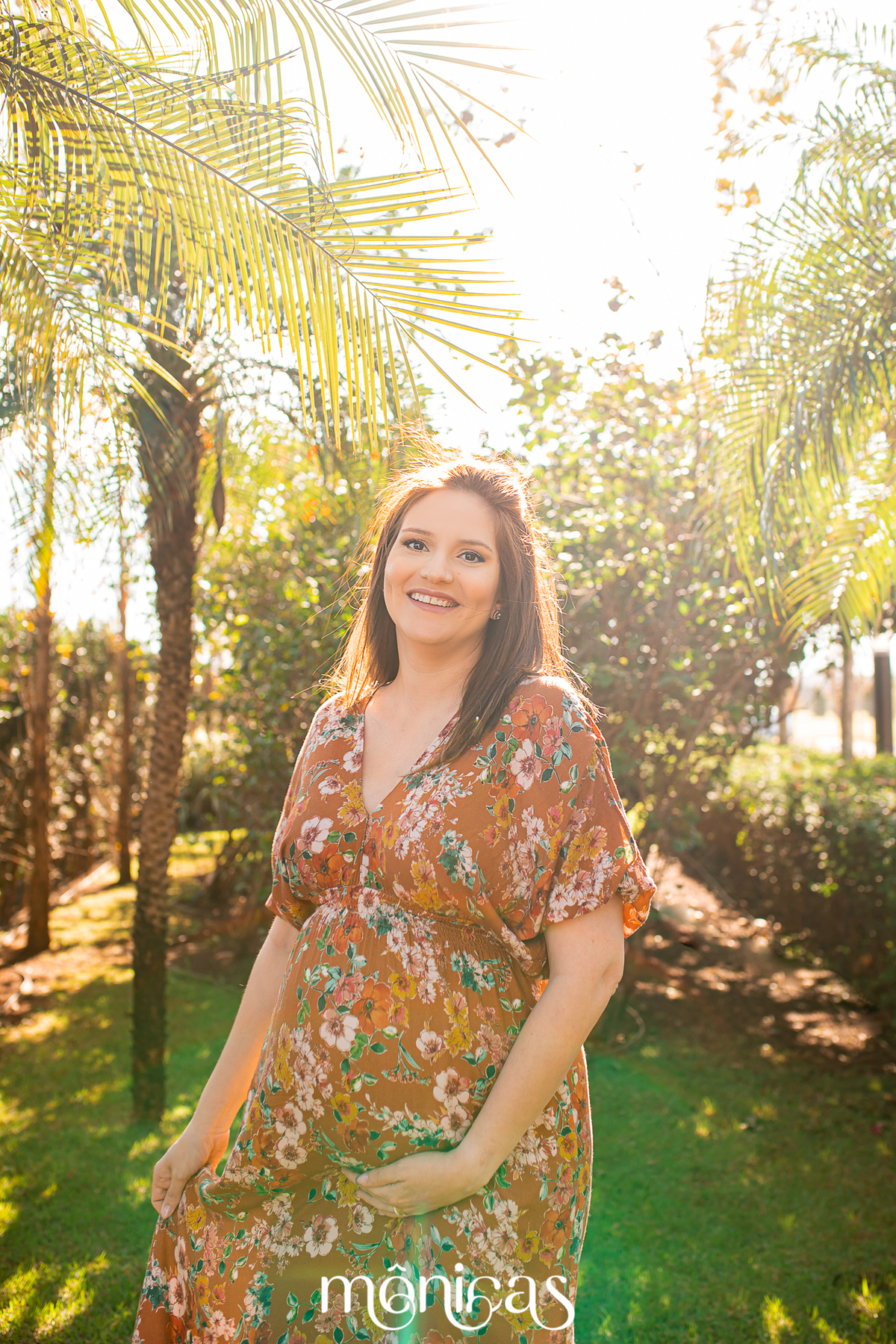 Caminhando sorrindo pelo parque, a gestante segura um vestido longo e olha para a fotógrafa
