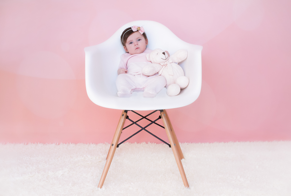 Em um fundo rosa, sentada numa cadeira branca, a Betina está segurando um ursinho de crochê