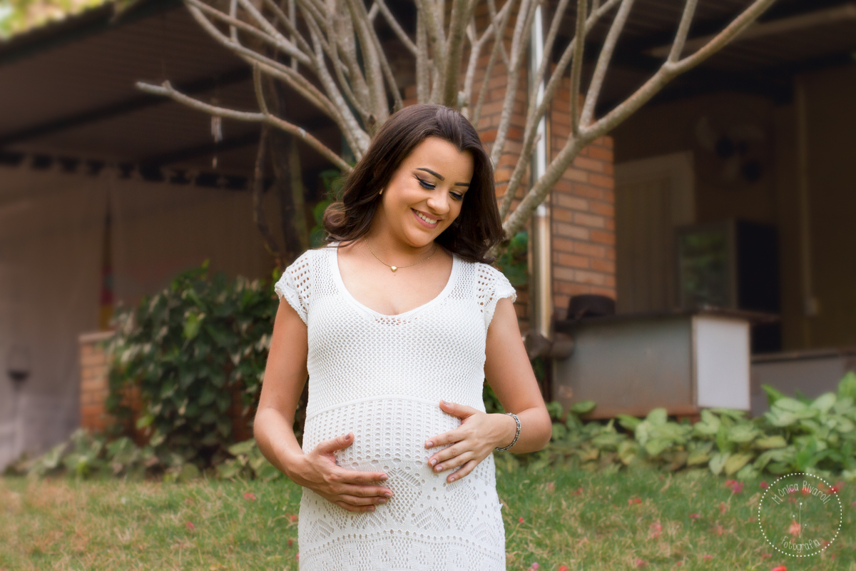 Em frente à uma árvore cheia de galhos, a Mari olha para sua barriga e sorri