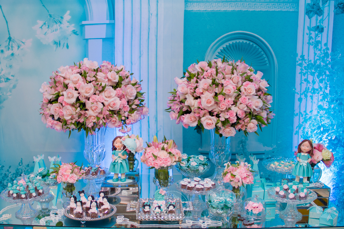 Close no lado esquerdo da mesa, também com doces, arranjo de rosas e a boneca inspirada na Lívia  