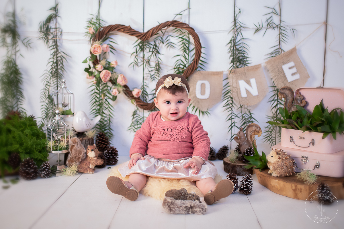 Em um cenário de bosque, a Maria Flor está sentadinha e sorri para a fotógrafa