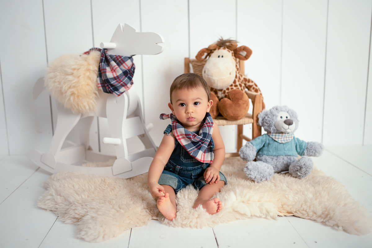 NEsta fotografia, a fotógrafa Mônica RIvaroli montou um cenário com pelúcias de urso, girafa e um cavalinho de madeira. O cavalinho usa uma bandana xadrez de azul, bege e vermelho igual à do Edu, que está sentadinho em meio aos bichinhos