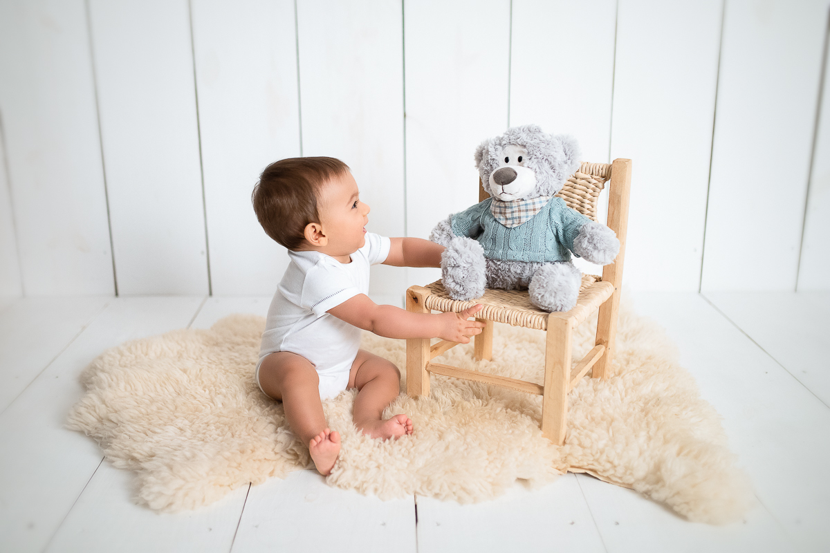 O Eduardo está sentadinho um pelinho de carneiro. Ao lado dele, a fotógrafa de bebês em Maringá/PR, colocou um ursinho em cima de uma cadeirinha de corda. O Edu olha para o ursinho e sorri